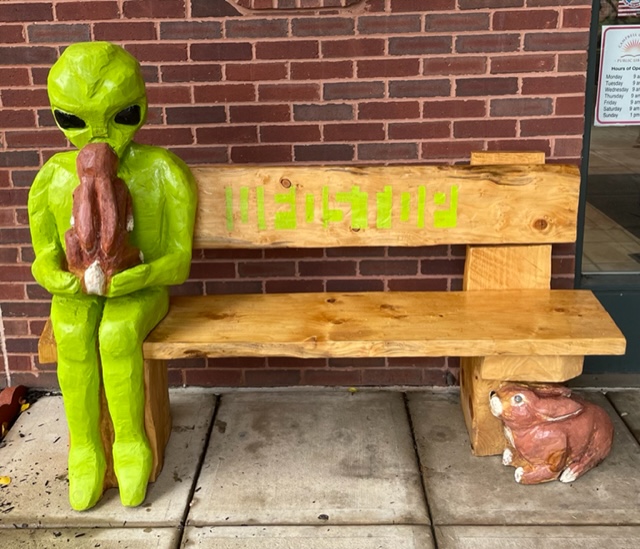 Depicted is a carved bench with an alien and bunny sitting on it, which was made by Chris Rust.